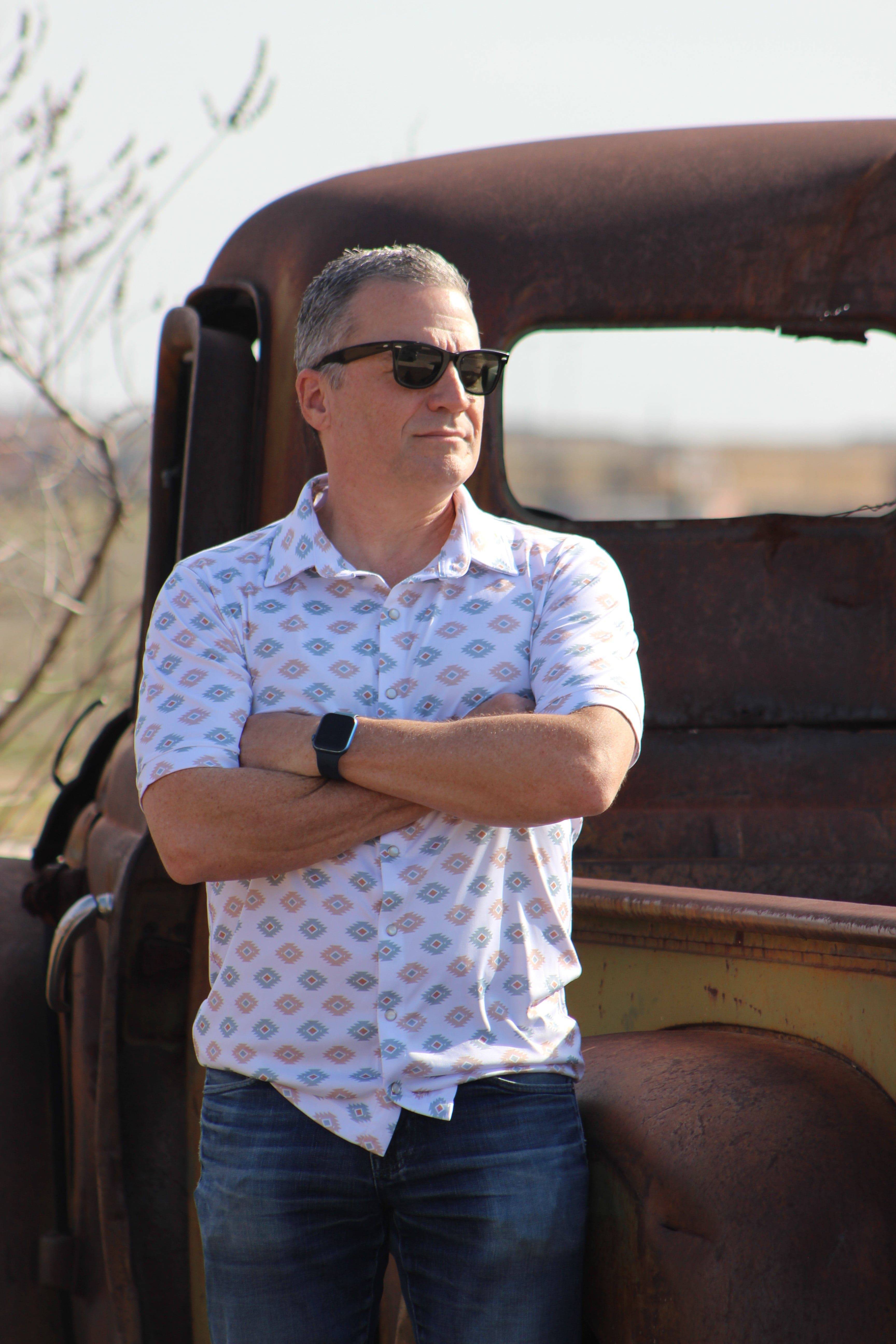 Confident man leaning against a rustic, vintage truck, wearing the 'Azure Earthshine' short-sleeved shirt from howdy-ho.com. The shirt's unique azure and beige pattern is a stylish contrast to the rugged backdrop. Accessorized with black sunglasses and a sleek smartwatch, he embodies a casual yet fashionable outdoor look. The image aims to showcase the shirt in a natural setting, illustrating its versatility and appeal for the modern man.