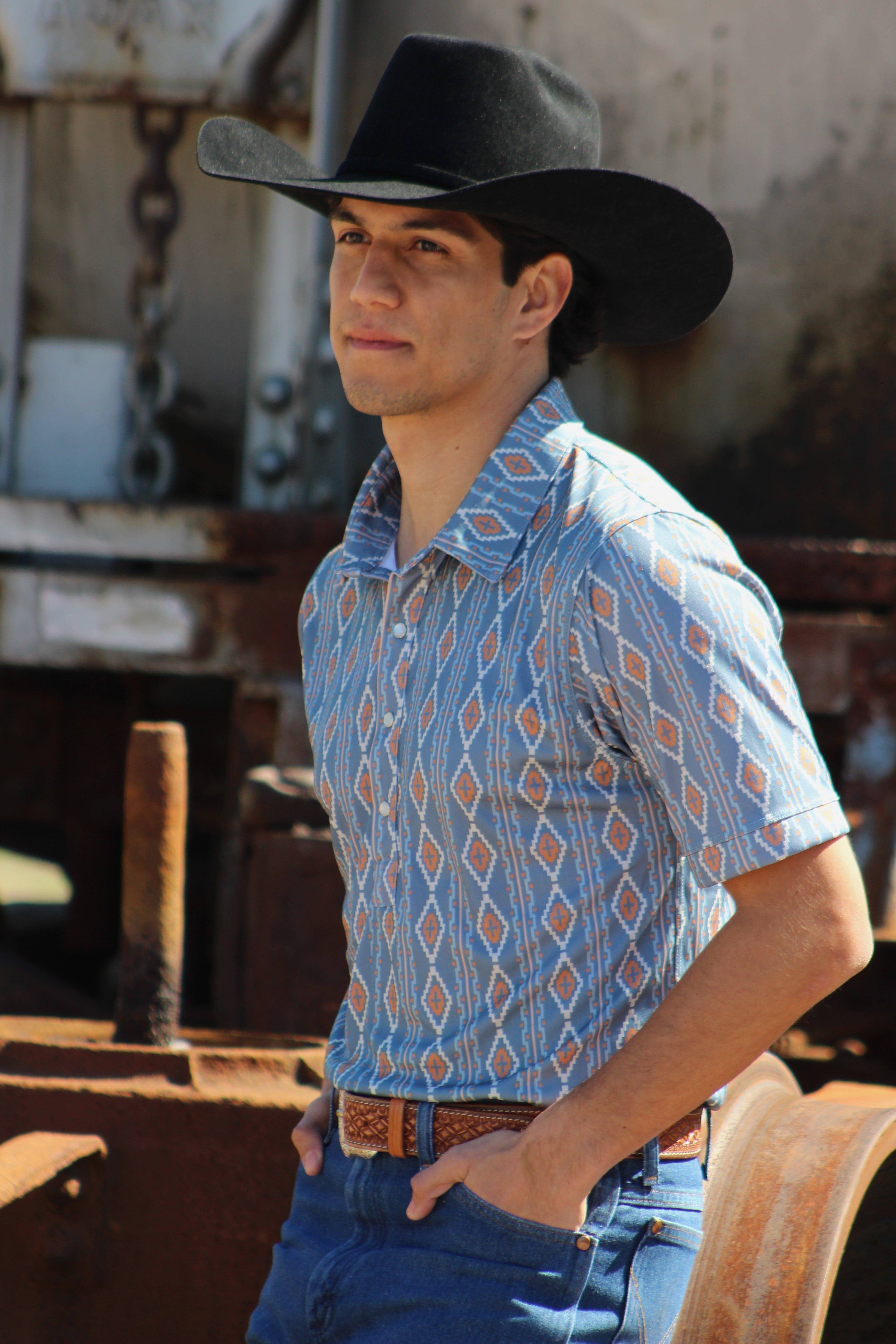 Confidently sporting the HowdyHo Aztec Brown Blue shirt, a man exudes Western charm with a modern twist. The shirt, featuring a distinctive Aztec pattern in shades of blue and brown, is perfectly complemented by a classic black cowboy hat, a leather belt, and blue jeans. He poses with a relaxed yet assertive attitude in an industrial setting, capturing the essence of HowdyHo's adventurous and masculine style.