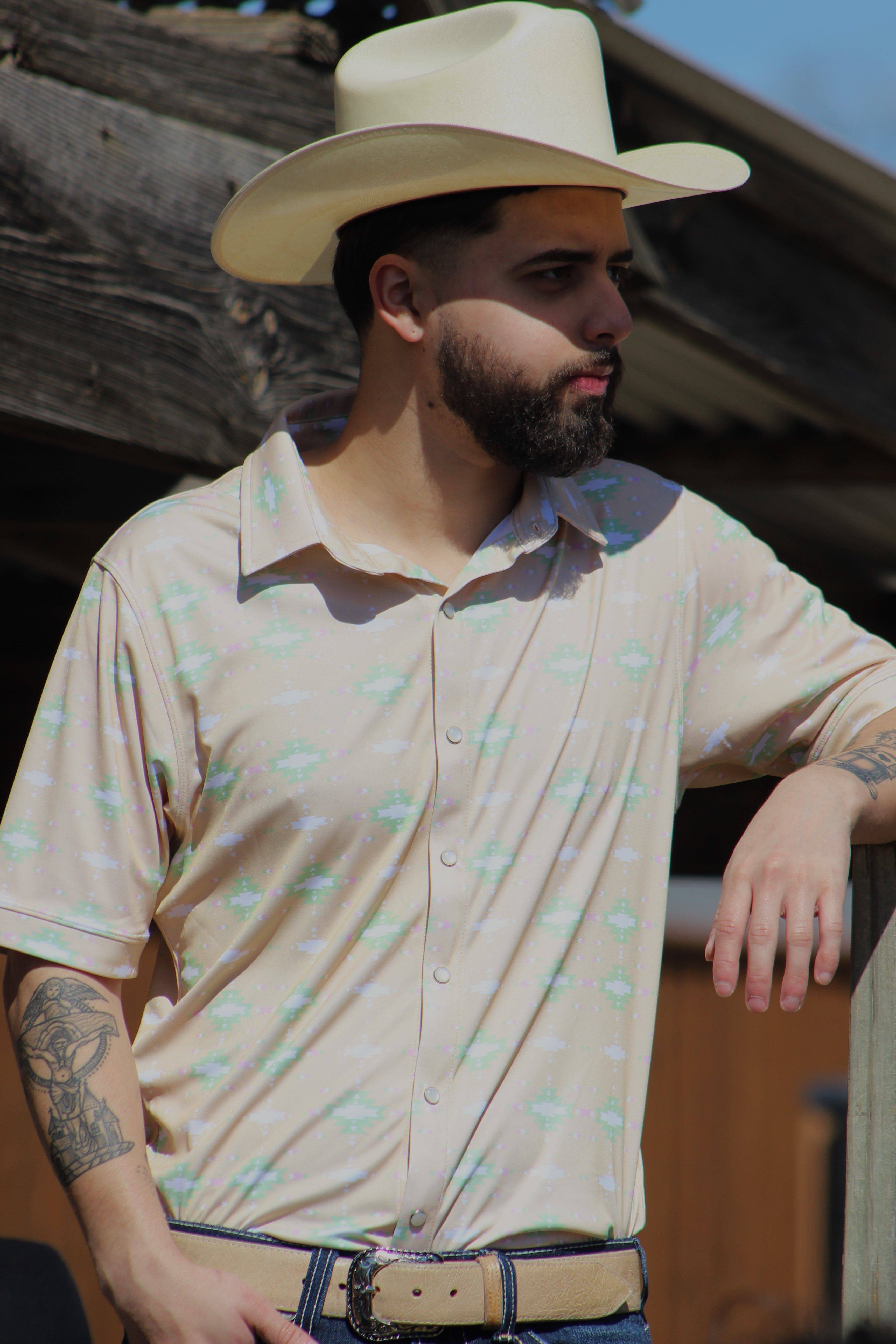 A stylish man leans confidently against a rustic wooden backdrop, wearing the Sunrise Serenity shirt from HowdyHo. The shirt's tan base is complemented by a subtle green and white floral design, perfect for a casual yet sophisticated look. Paired with a classic beige cowboy hat, dark jeans, and a detailed leather belt, this ensemble captures the essence of modern western style.