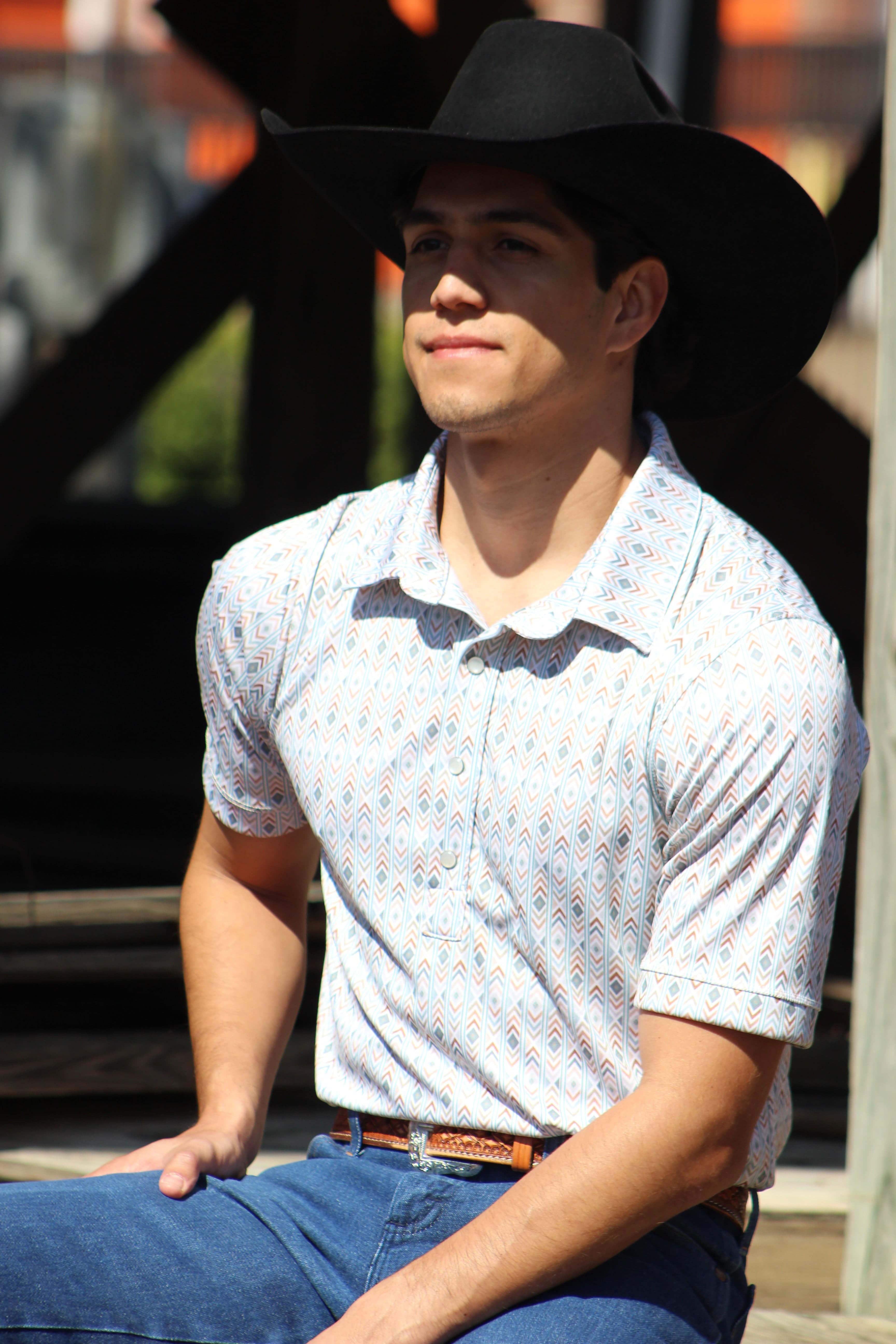 A confident man gazes into the distance, wearing HowdyHo's Aztec Fusion print shirt topped with a classic black cowboy hat. The shirt's pattern showcases a playful mix of geometric shapes in soft blues and whites, perfectly paired with dark blue jeans and a detailed leather belt, capturing the spirit of the modern West.