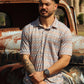 A man stands in front of a vintage rusted truck, modeling the HowdyHo Aztec Twilight Blend shirt. The short-sleeved shirt boasts a complex Aztec pattern in shades of blue and orange, offering a modern twist on classic southwestern motifs. His casual stance, with arms crossed, along with the rugged outdoor backdrop, highlights the shirt's adventurous spirit and durability.