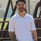 A man with a warm smile is casually leaning on a wooden barrel, wearing the Sunset Crossroads shirt from HowdyHo. The shirt boasts a soft pattern of yellow suns and pink crosses on a light background, creating a fun and fresh look. His relaxed pose, paired with jeans and a stylish watch, captures the essence of HowdyHo's blend of comfort and trendy design.
