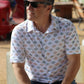 Confident man casually seated outdoors, sporting the 'Azure Earthshine' short-sleeved shirt from howdy-ho.com. The shirt's azure and beige geometric pattern pairs well with his dark denim jeans, complemented by classic black sunglasses and a smartwatch. This lifestyle image captures the shirt's relaxed fit and stylish design, ideal for a sunny day out, giving potential customers a real-world look at the product.
