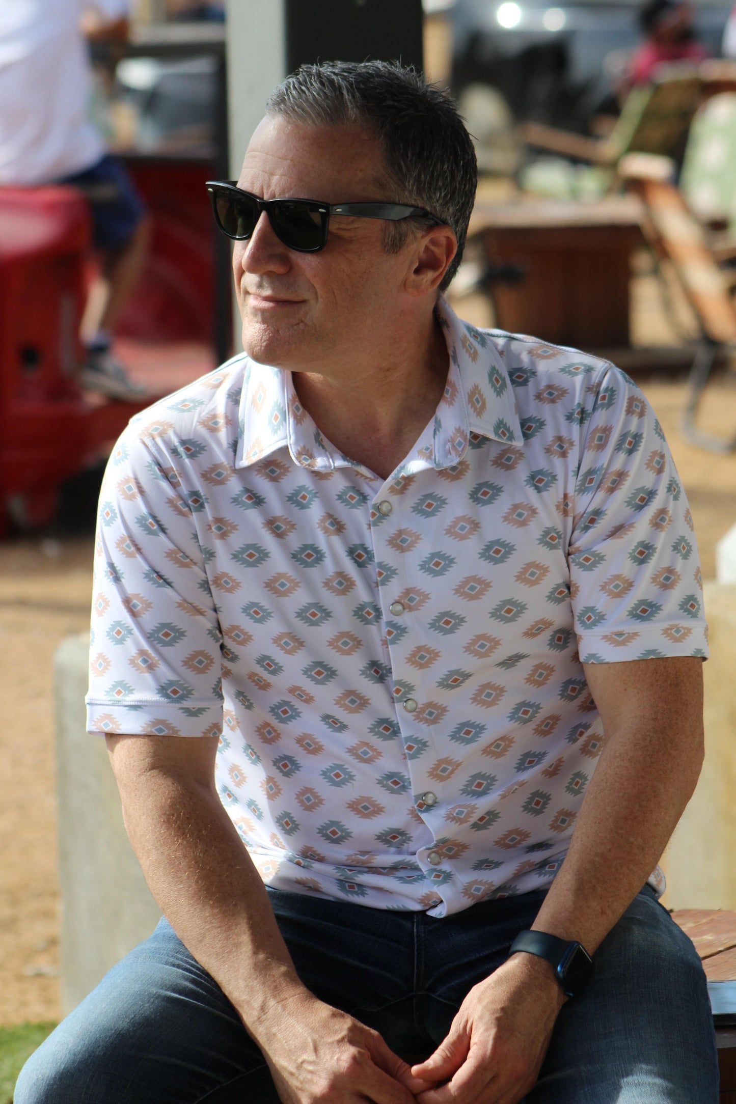 Confident man casually seated outdoors, sporting the 'Azure Earthshine' short-sleeved shirt from howdy-ho.com. The shirt's azure and beige geometric pattern pairs well with his dark denim jeans, complemented by classic black sunglasses and a smartwatch. This lifestyle image captures the shirt's relaxed fit and stylish design, ideal for a sunny day out, giving potential customers a real-world look at the product.