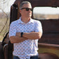 Confident man leaning against a rustic, vintage truck, wearing the 'Azure Earthshine' short-sleeved shirt from howdy-ho.com. The shirt's unique azure and beige pattern is a stylish contrast to the rugged backdrop. Accessorized with black sunglasses and a sleek smartwatch, he embodies a casual yet fashionable outdoor look. The image aims to showcase the shirt in a natural setting, illustrating its versatility and appeal for the modern man.