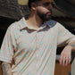 A stylish man leans confidently against a rustic wooden backdrop, wearing the Sunrise Serenity shirt from HowdyHo. The shirt's tan base is complemented by a subtle green and white floral design, perfect for a casual yet sophisticated look. Paired with a classic beige cowboy hat, dark jeans, and a detailed leather belt, this ensemble captures the essence of modern western style.
